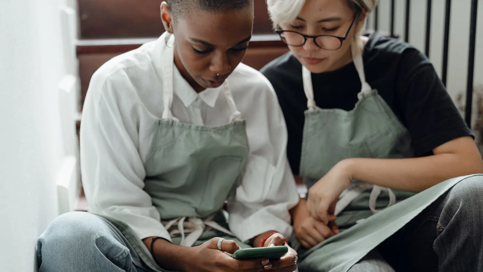 Two coworkers in aprons sit on a staircase during a break, looking at a lesson on a smartphone