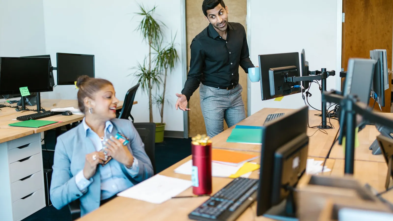Two colleagues chat and smile beside a desk in a bright office