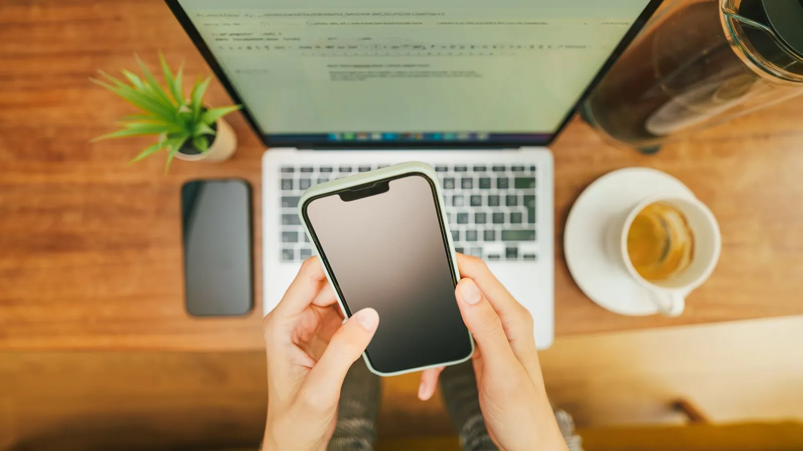 Hands hold a smartphone above a laptop on a wooden desk, next to a cup of coffee